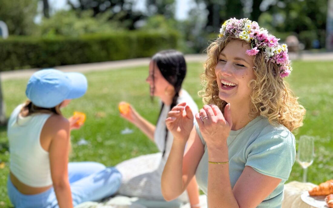 Eine junge Frau mit lockigem Haar und Blumenkranz lächelt fröhlich bei einem Midsommar-Picknick. Hinter ihr sitzen zwei Freundinnen im Gras, die an einem sonnigen Tag Getränke reichen.