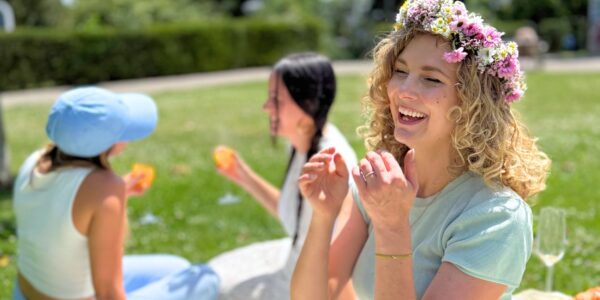 Eine junge Frau mit lockigem Haar und Blumenkranz lächelt fröhlich bei einem Midsommar-Picknick. Hinter ihr sitzen zwei Freundinnen im Gras, die an einem sonnigen Tag Getränke reichen.